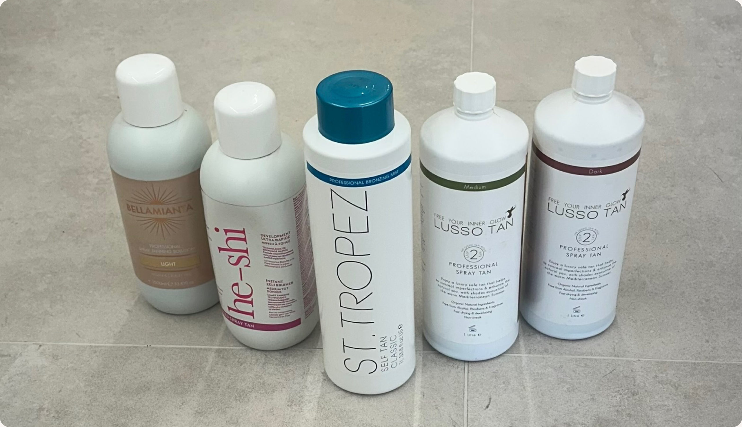 Professional spray tanning solution bottles including Bellamianta, He-Shi, St. Tropez, and Lusso Tan arranged on a tiled floor